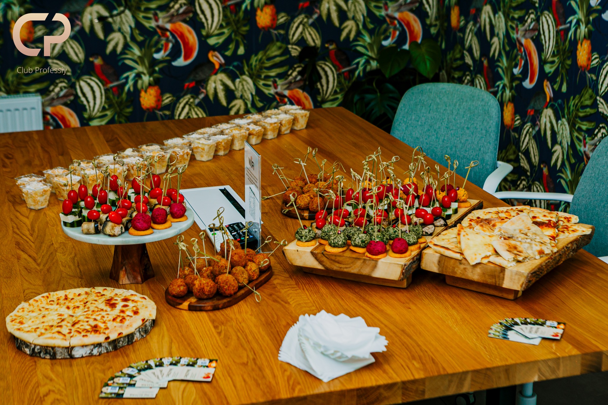 Stół cateringowy z przekąskami: koreczki z pomidorkami, kulki mięsne, pizza na drewnianej desce, sałatki w kubeczkach. W tle krzesła i ściana w tropikalne wzory.