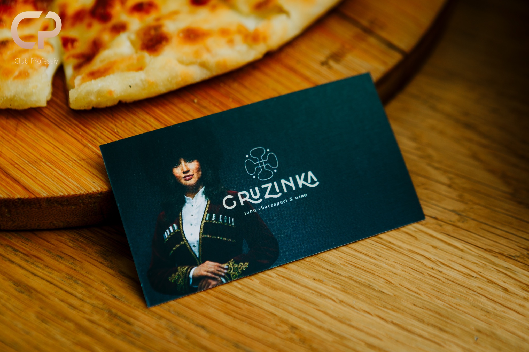 Karta wizytowa restauracji Gruzinka leży na drewnianym blacie obok kawałka chaczapuri. Karta prezentuje wizerunek kobiety w tradycyjnym stroju gruzińskim.
