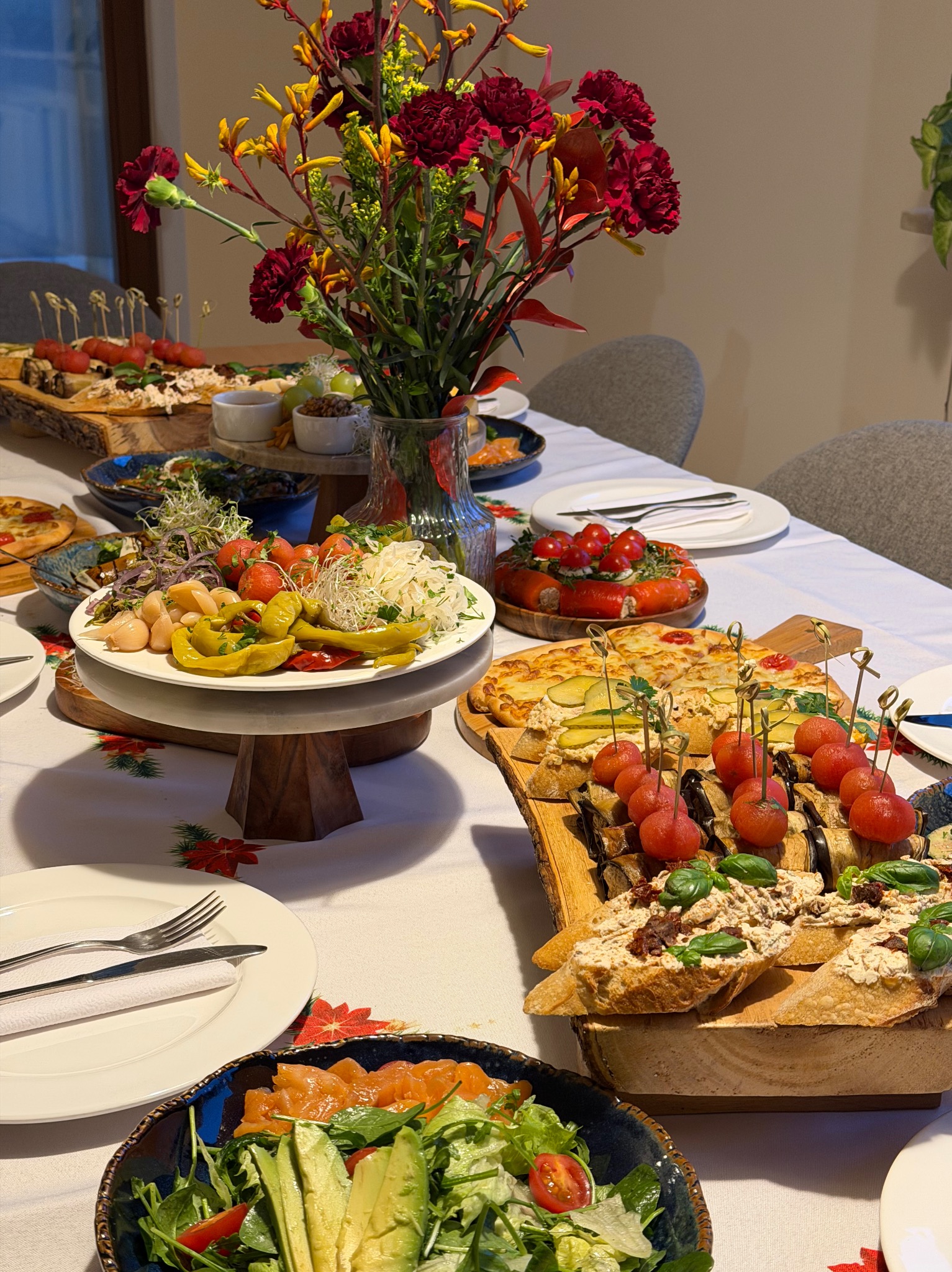 Stół z bogato zastawionymi potrawami: sałatki, przekąski na drewnianych deskach, kwiaty w wazonie. Dekoracja stołu z motywem świątecznym.