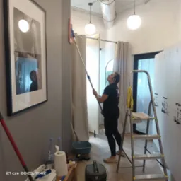 Pracownica w czarnym stroju myje ścianę mopem teleskopowym w nowoczesnym wnętrzu. Widoczna drabina, akcesoria do sprzątania i dekoracyjny obraz na ścianie.