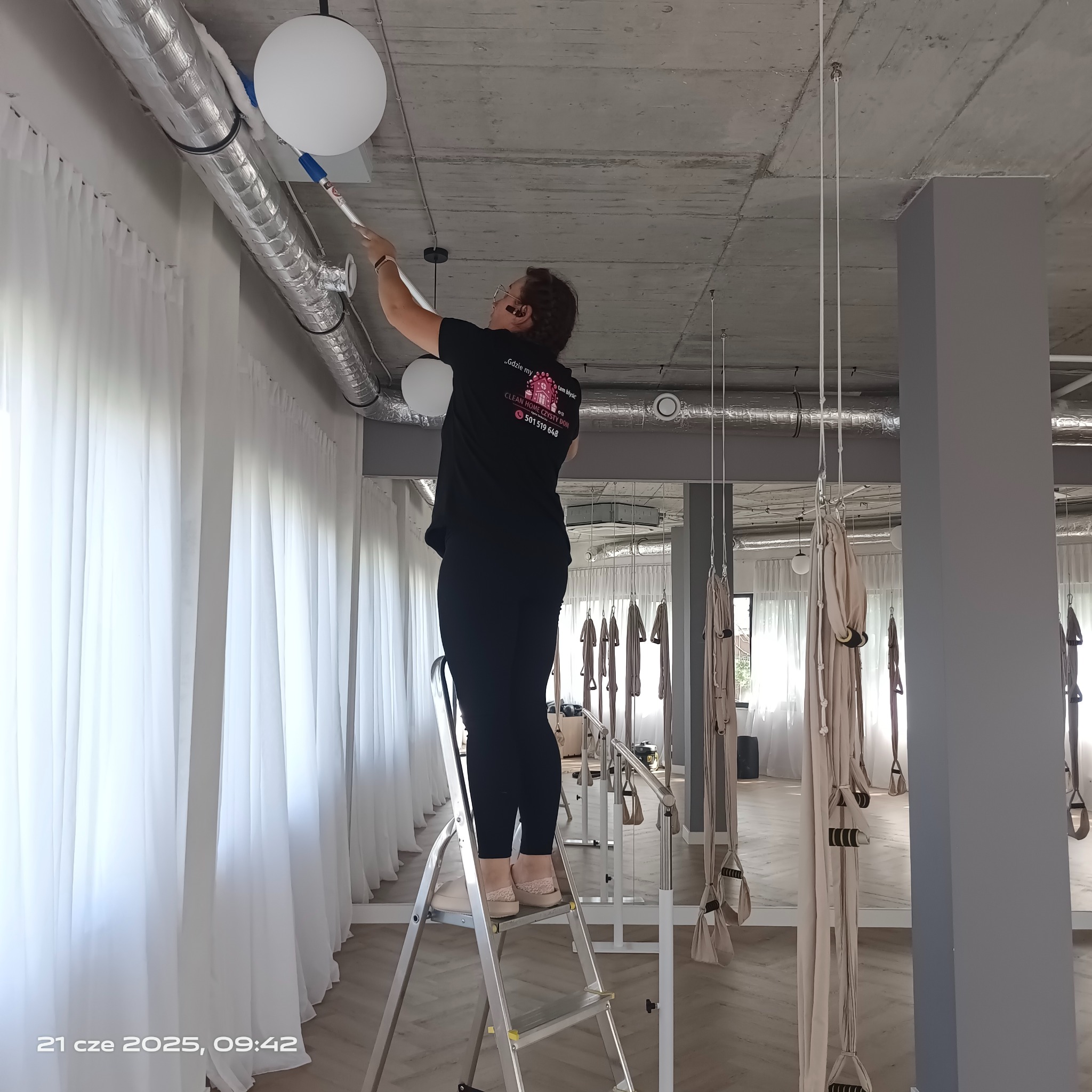 Kobieta na drabinie czyści białą lampę sufitową w pomieszczeniu z elementami do aerial jogi. Widoczne rury wentylacyjne pod sufitem i białe zasłony.