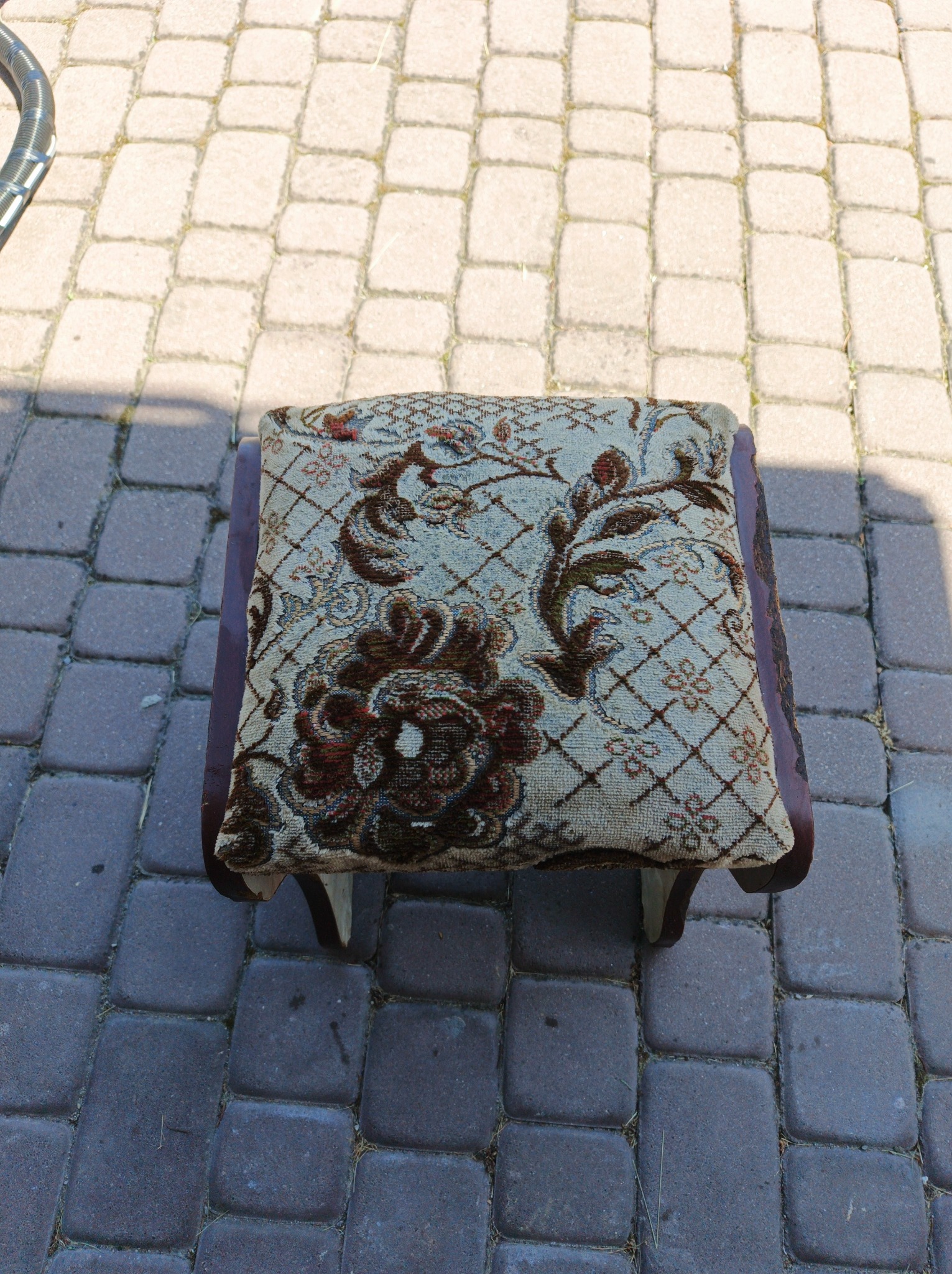 Tapicerowany puf z wzorem kwiatowym na kostce brukowej, widok z góry. Fragment węża odkurzacza w lewym górnym rogu sugeruje pranie tapicerki.