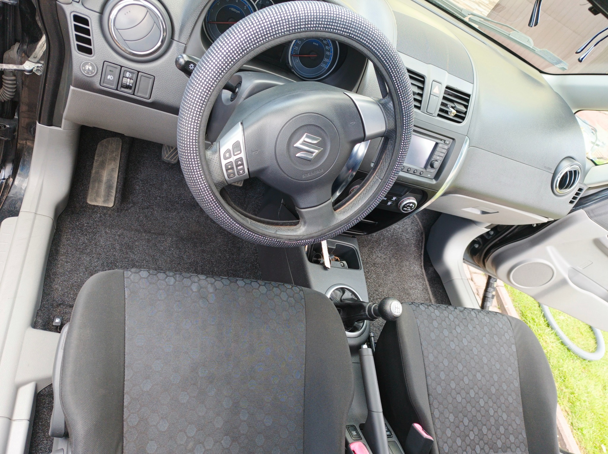 Wnętrze Suzuki po praniu: czysta tapicerka foteli z geometrycznym wzorem, kierownica z pokrowcem w kropki, dywaniki bez plam. Widoczna dźwignia zmiany biegów i deska rozdzielcza.