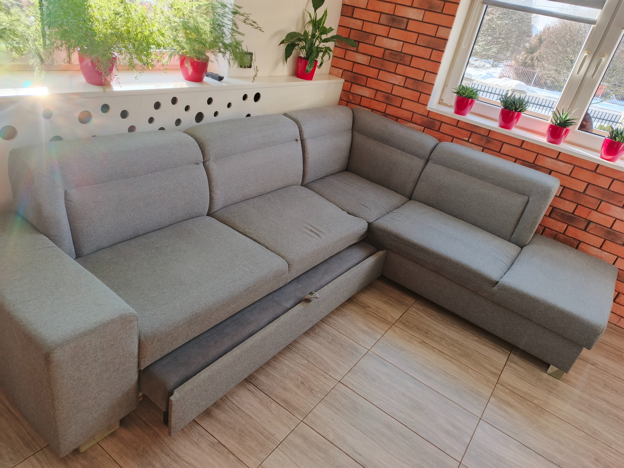 Szara, rozłożona kanapa narożna z wysuniętą szufladą na pościel, na tle ceglanej ściany i okna z roślinami w czerwonych doniczkach. Widok z góry.