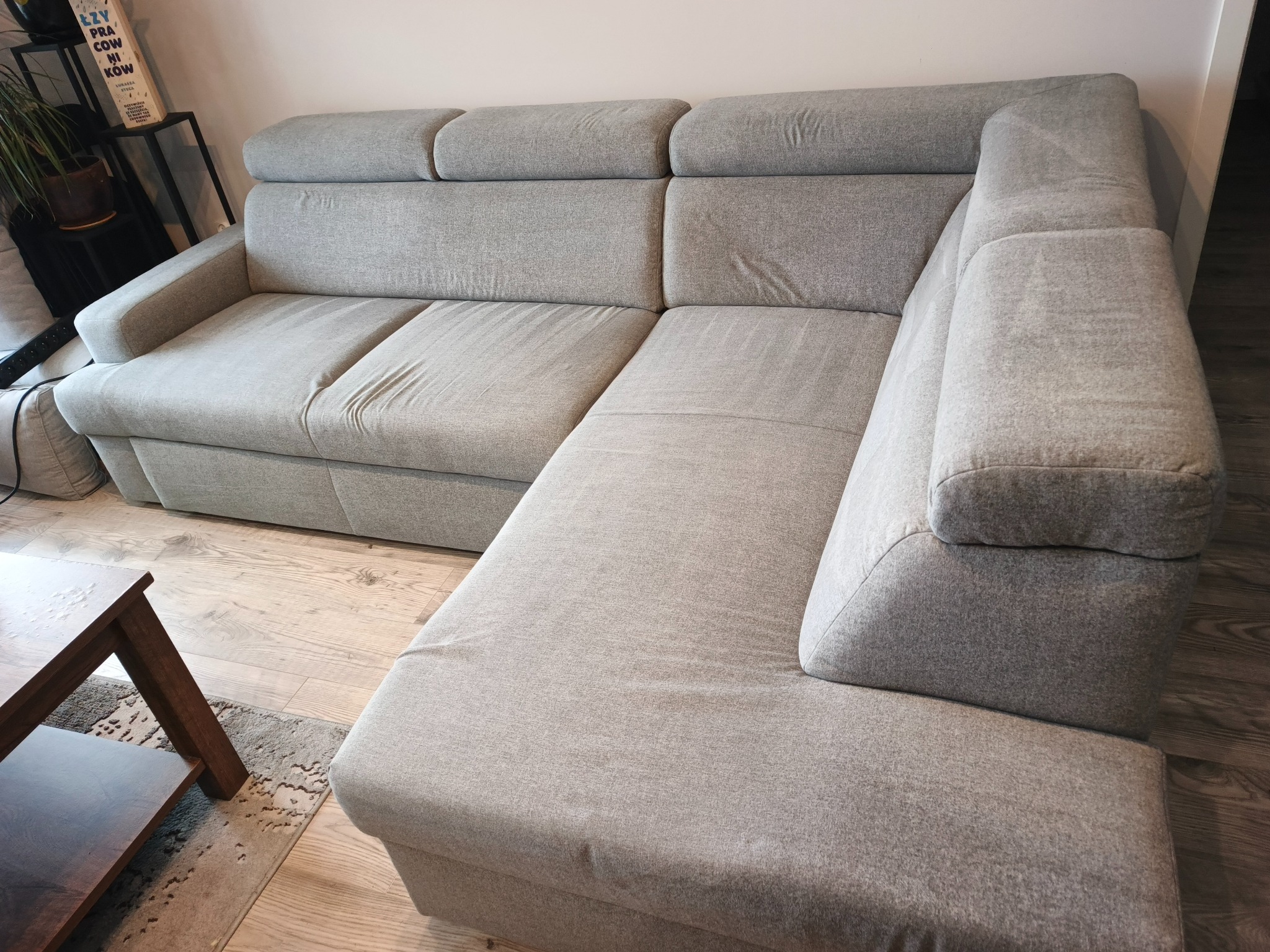 Jasnoszara, narożna kanapa z regulowanymi zagłówkami, ustawiona na drewnianej podłodze. Widoczny fragment stolika kawowego i regału z książkami w tle.