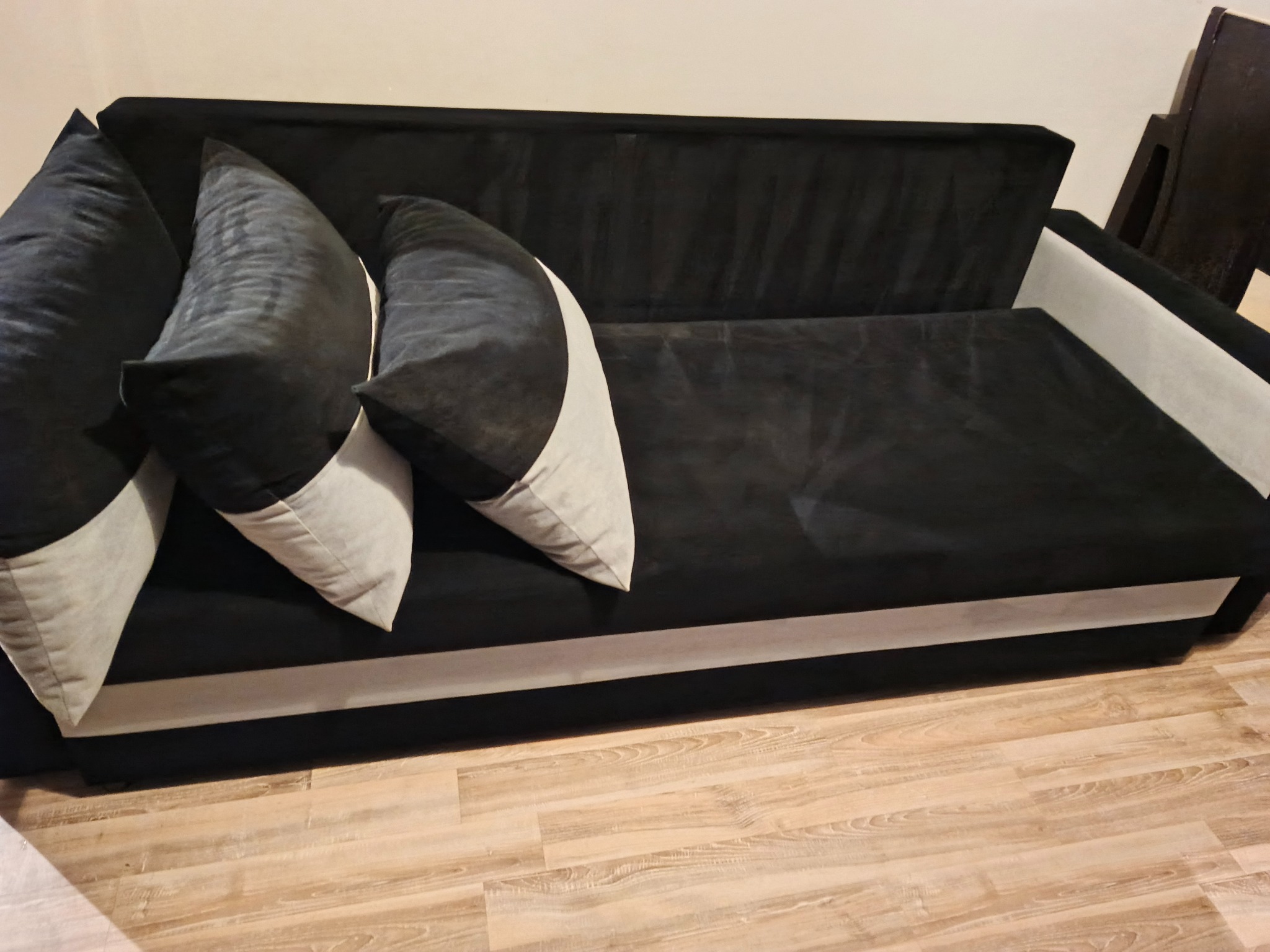 Czarno-szara rozkładana kanapa z trzema poduszkami na drewnianej podłodze. Widok z boku, oświetlenie naturalne.