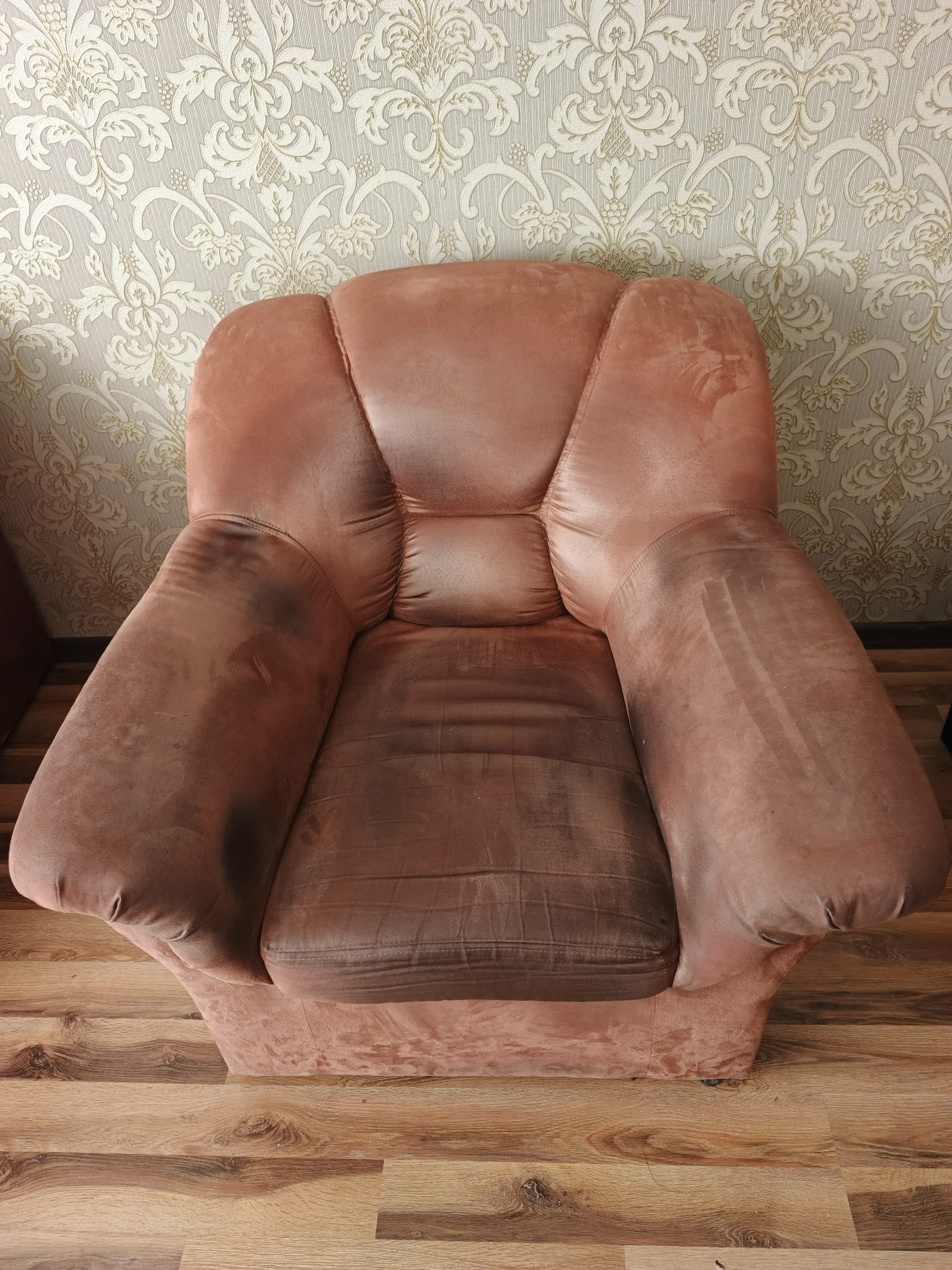 Brudny, pluszowy fotel w odcieniach brązu i różu, z widocznymi plamami na siedzisku i podłokietnikach, na tle wzorzystej tapety i drewnianej podłogi.
