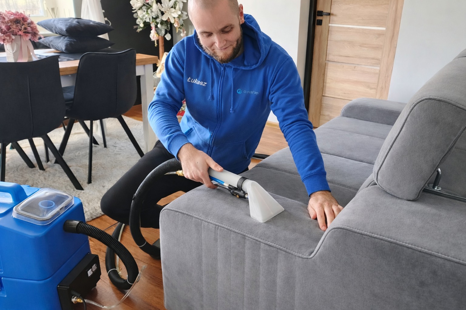 Mężczyzna w niebieskiej bluzie czyści szarą kanapę za pomocą profesjonalnego sprzętu do prania tapicerki w jasnym wnętrzu. Widoczny sprzęt i detale kanapy.