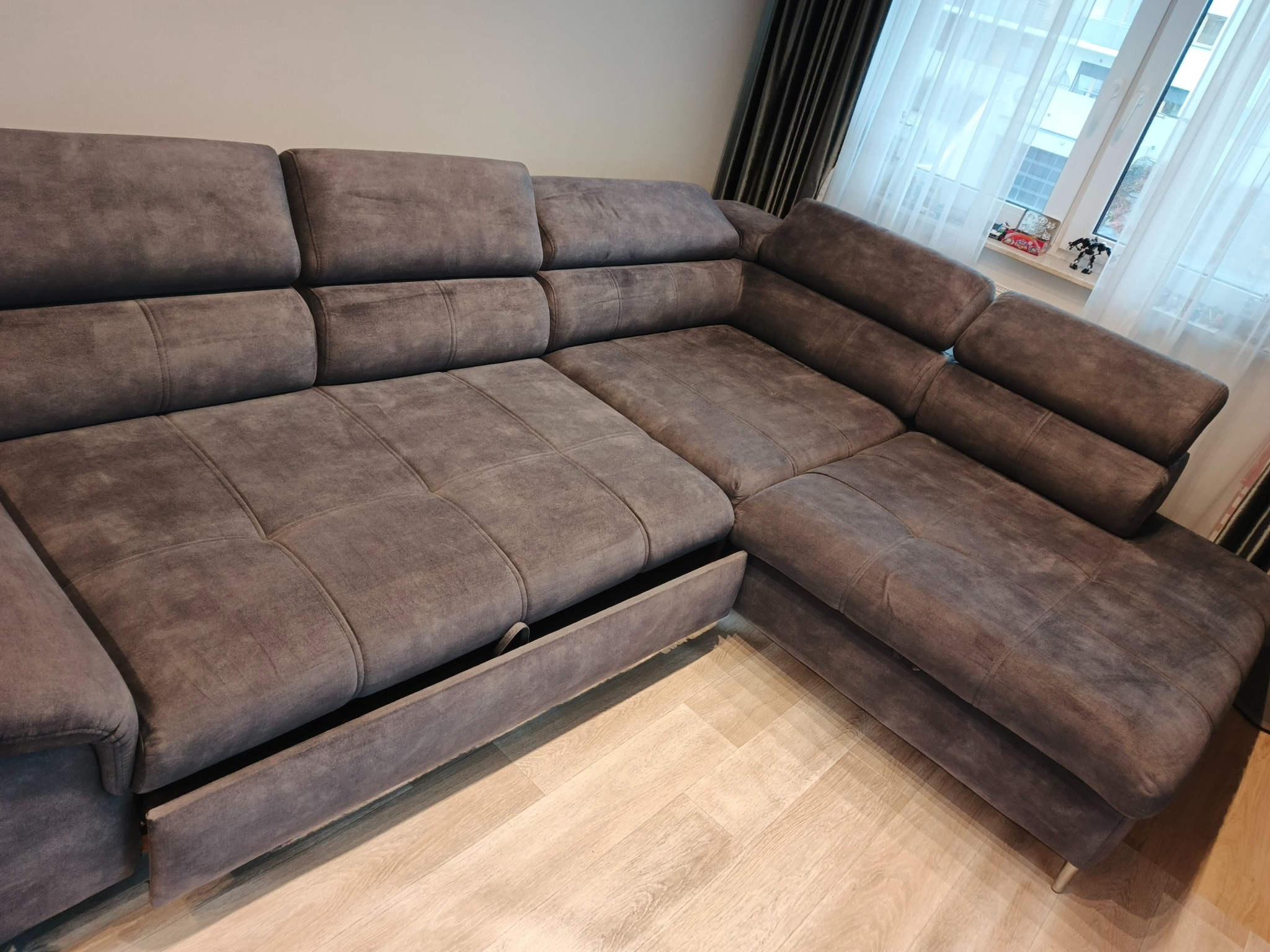 Szara, narożna kanapa z wysuwaną szufladą na pościel, ustawiona na jasnej podłodze, z oknem w tle. Widoczne detale tapicerki i metalowe nóżki.