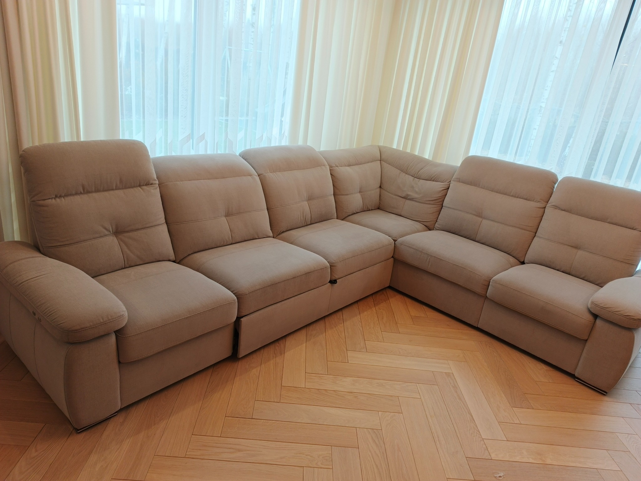 Beżowy narożnik z funkcją spania na parkiecie w jodełkę, oświetlony naturalnym światłem wpadającym przez okno z białymi zasłonami. Widok z przodu, lekko z góry.