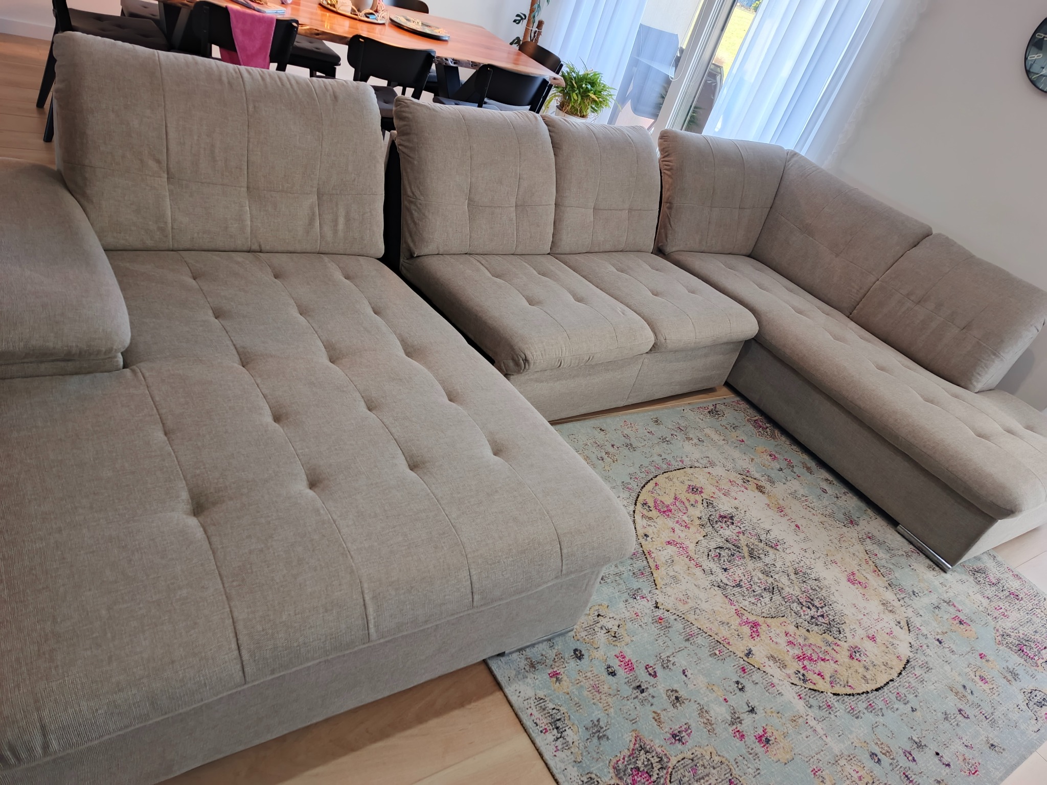 Jasna, narożna kanapa z tapicerowanymi guzikami na siedzisku, ustawiona na wzorzystym dywanie w salonie. Widok z góry, naturalne światło.