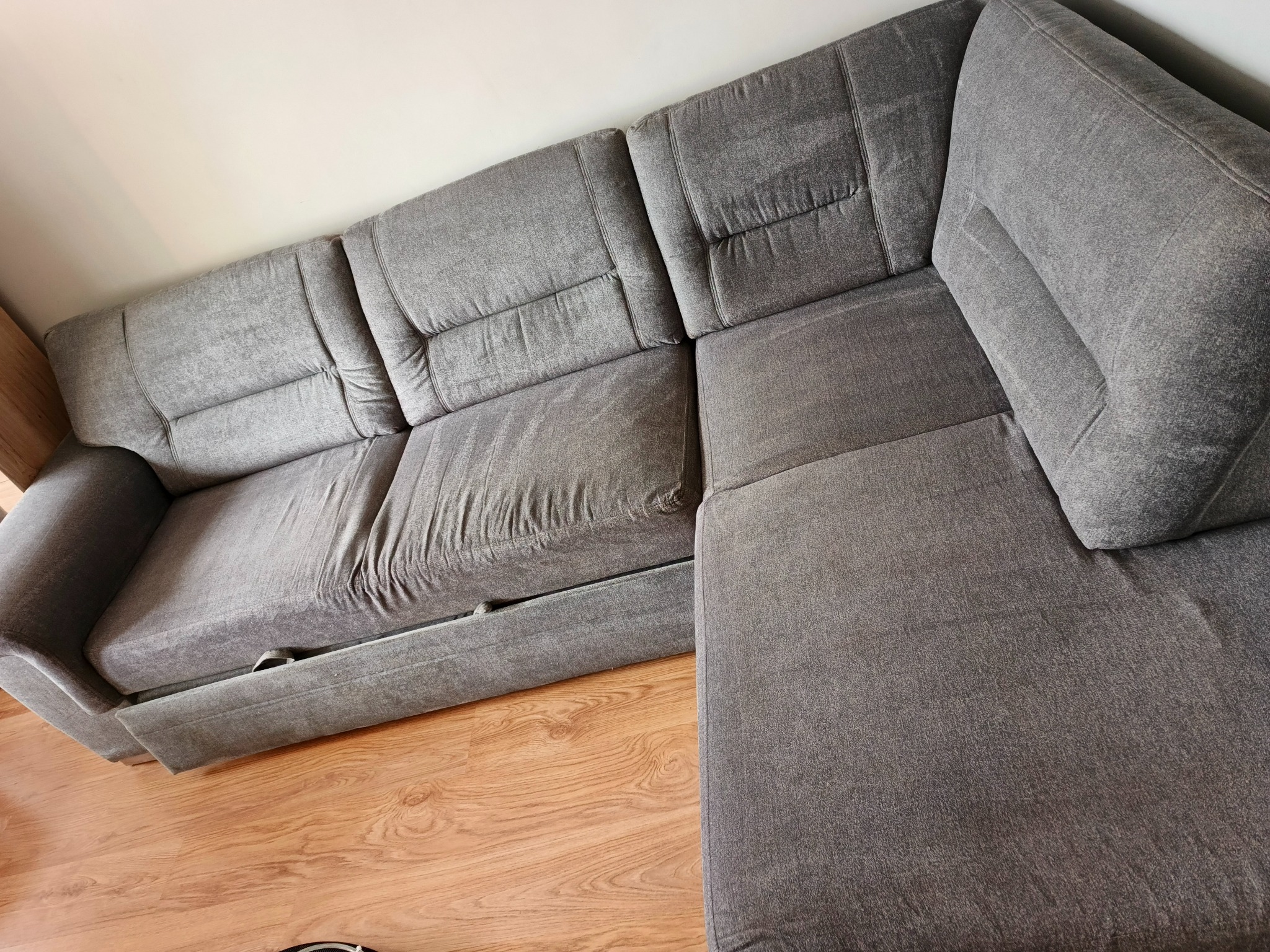 Szara, narożna kanapa z welurową tapicerką, widoczna z góry pod kątem, na drewnianej podłodze. Widoczne detale przeszyć i faktury materiału.