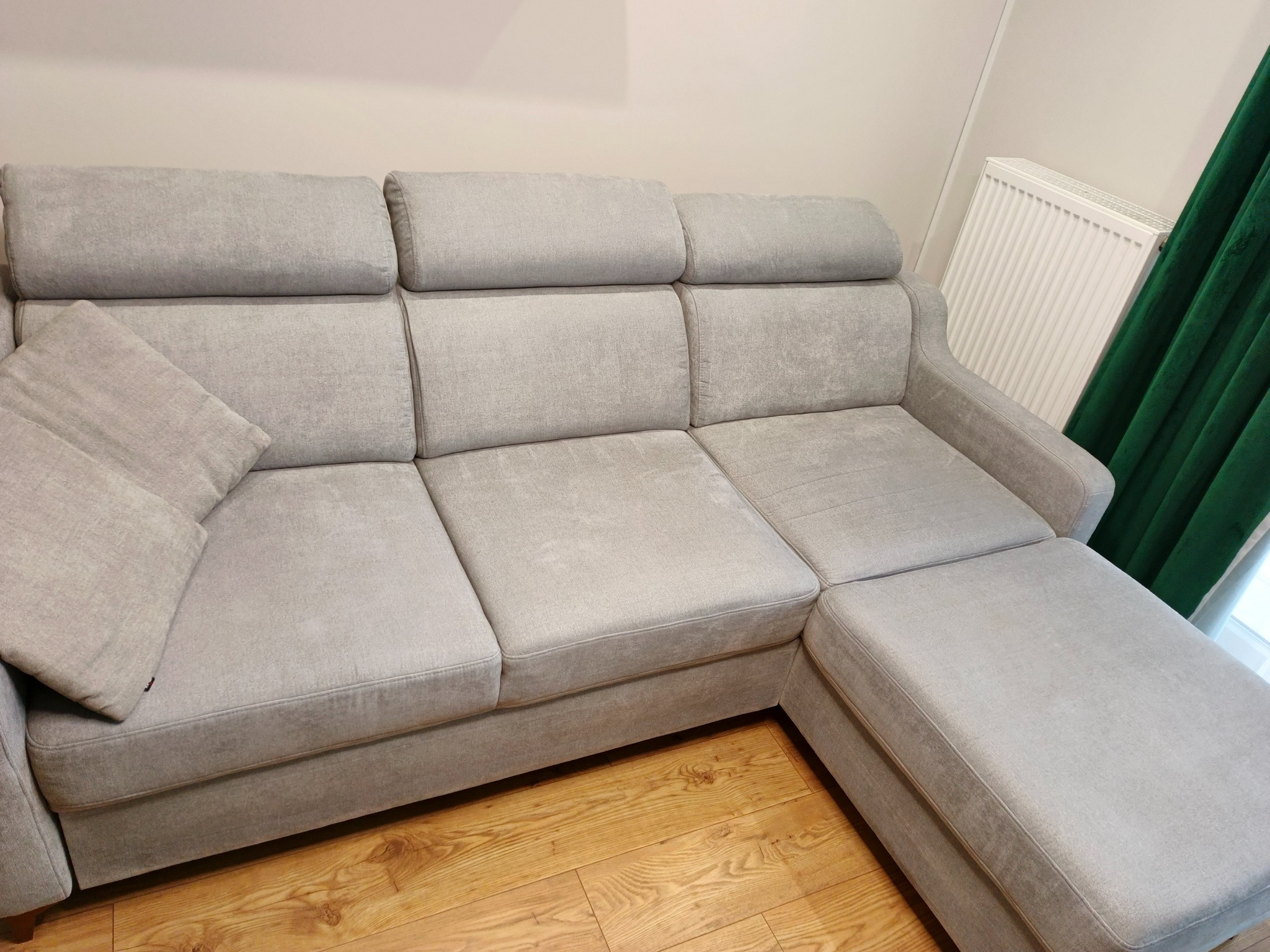 Jasnoszara, rozłożysta kanapa z poduszką, widoczna na tle drewnianej podłogi i fragmentu zielonej zasłony. Ujęcie z perspektywy lekko z góry, podkreślające fakturę materiału.