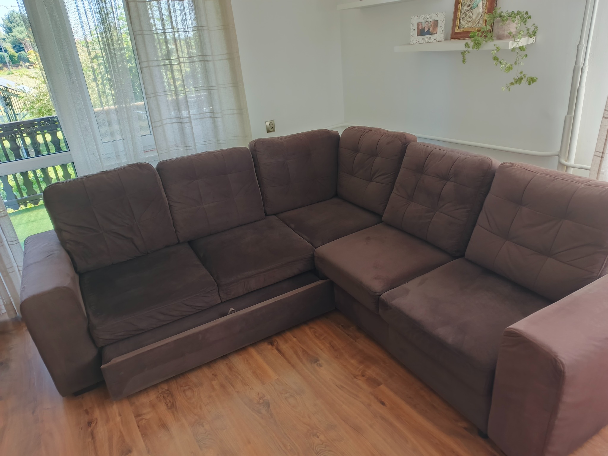 Ciemnobrązowa, narożna kanapa z wysuwaną szufladą na drewnianej podłodze. W tle okno z widokiem na zieleń i biała ściana z półką i dekoracjami.