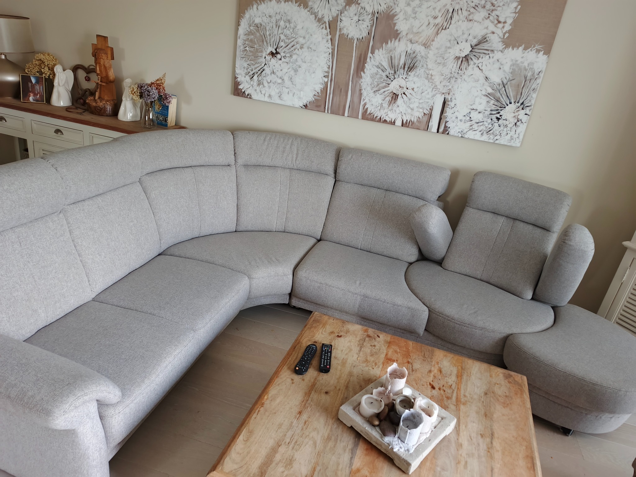 Jasnoszara, modułowa kanapa narożna z podnóżkiem, drewniany stolik kawowy z dekoracjami, obraz z dmuchawcami na ścianie. Widok na salon.