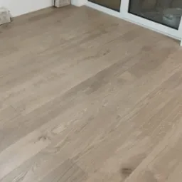 Jasne panele podłogowe ułożone w pokoju, widoczne puszki po panelach przy ścianie i okno balkonowe. Nowa podłoga po montażu.