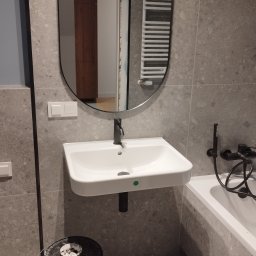 Siergiej Garamiew - Nowoczesna łazienka we Wrocławiu z umywalką na tle szarych płytek i okrągłego lustra. Widoczne wiaderka z farbą, sugerujące świeżo zakończone prace malarskie.