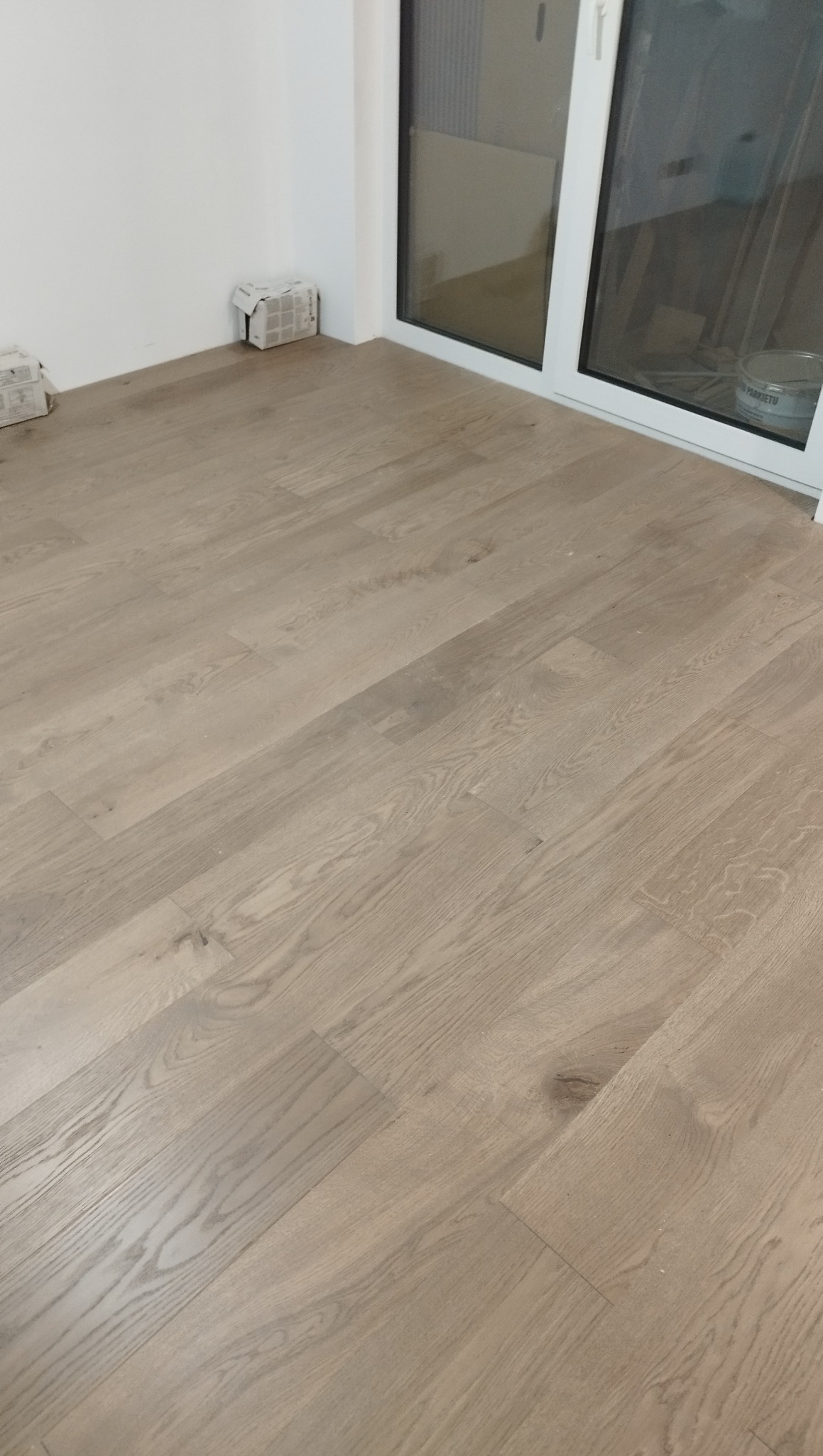 Jasne panele podłogowe ułożone w pokoju, widoczne puszki po panelach przy ścianie i okno balkonowe. Nowa podłoga po montażu.