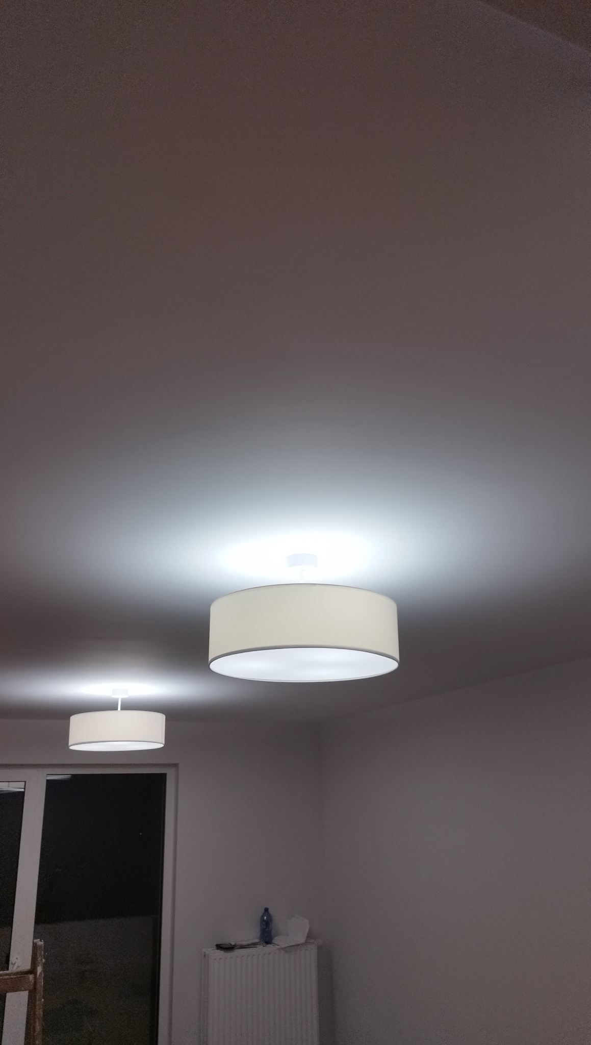 Dwa okrągłe, podwieszane plafony z materiałowym abażurem oświetlają wnętrze pokoju po malowaniu. Widoczny grzejnik i okno.