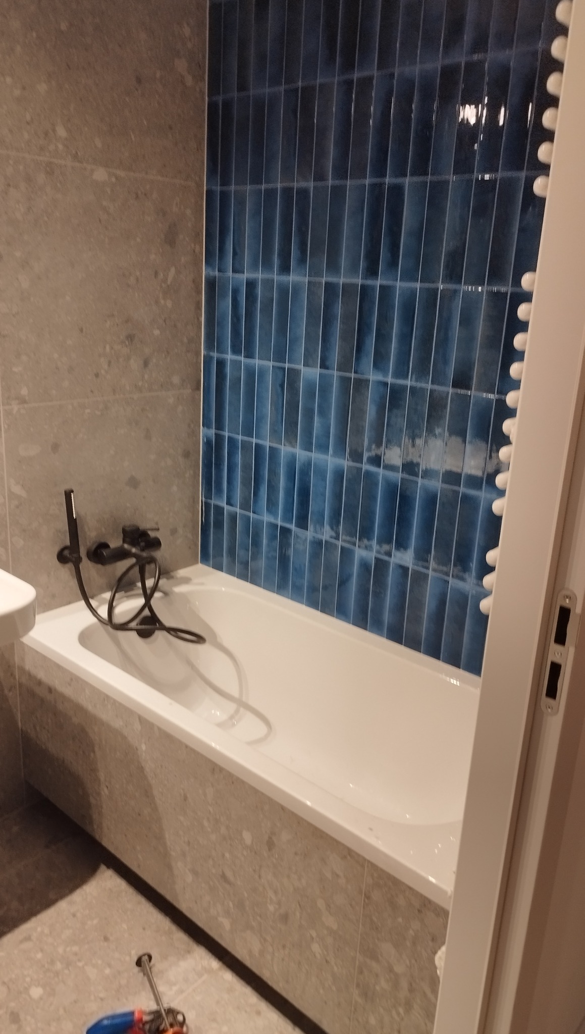 Wykończona łazienka z białą wanną, czarną armaturą i ścianą w niebieskich, pionowych płytkach. Widoczne narzędzia na podłodze.