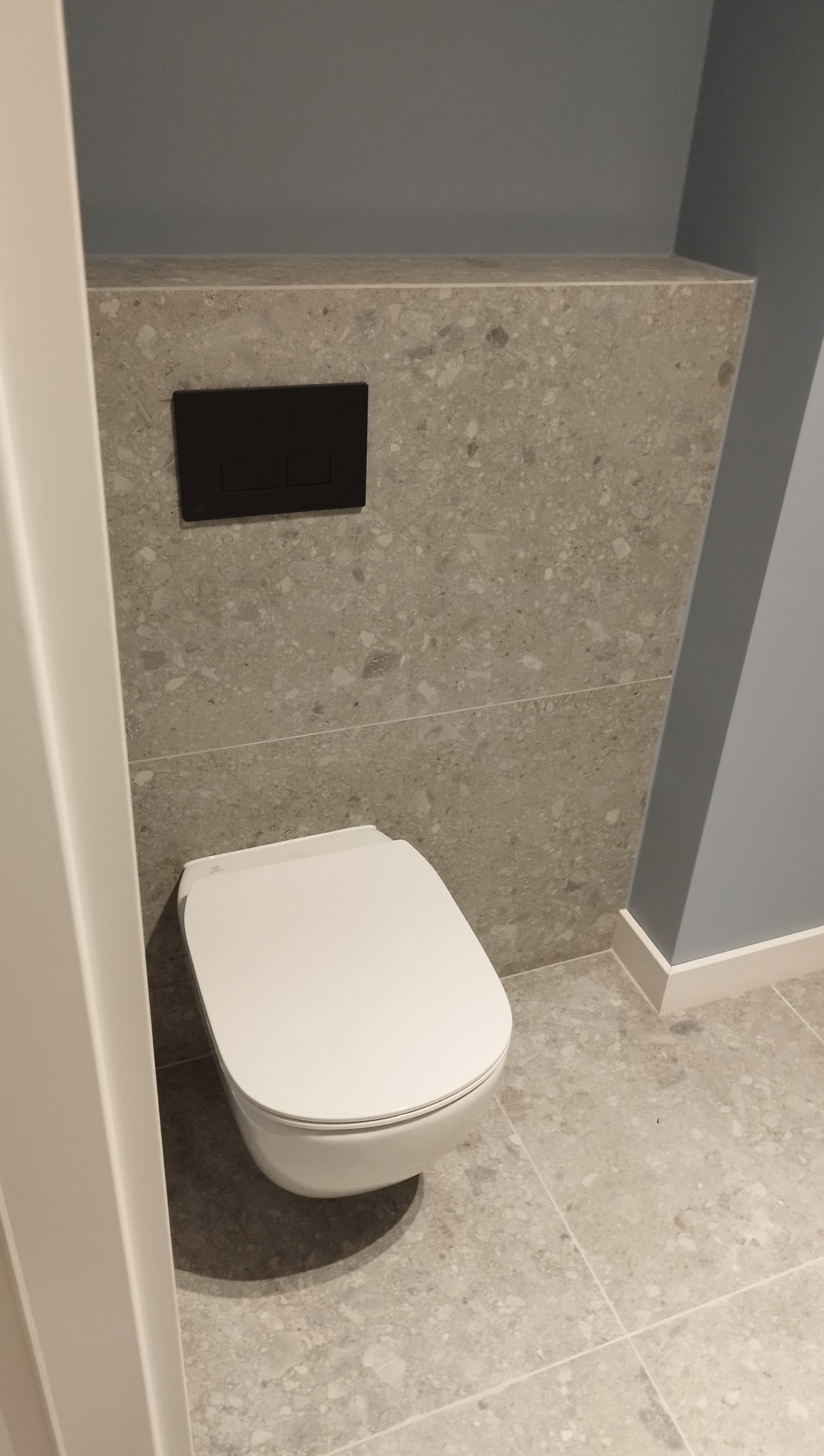 Nowoczesna toaleta podwieszana z czarnym panelem spłukującym na tle ściany wykończonej płytkami imitującymi lastryko i szarej ściany. Minimalistyczny design.