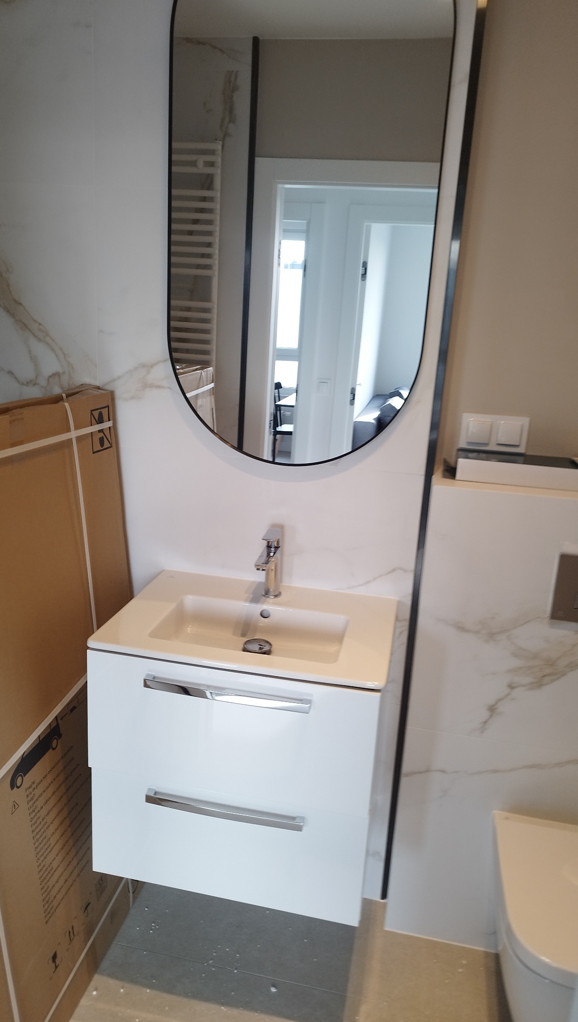 Nowoczesna biała szafka umywalkowa z chromowanymi uchwytami, owalne lustro z czarną ramą, marmurowe płytki na ścianie i podłodze, widok na dalsze pomieszczenie.