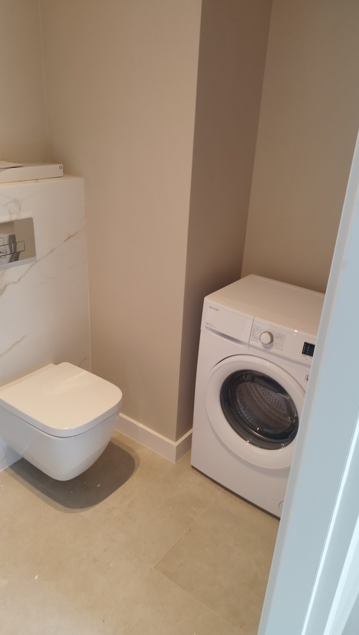 Jasna łazienka po remoncie: podwieszana toaleta z marmurowym wzorem, beżowe ściany i biała pralka. Minimalistyczny styl, świeżo po odnowieniu.