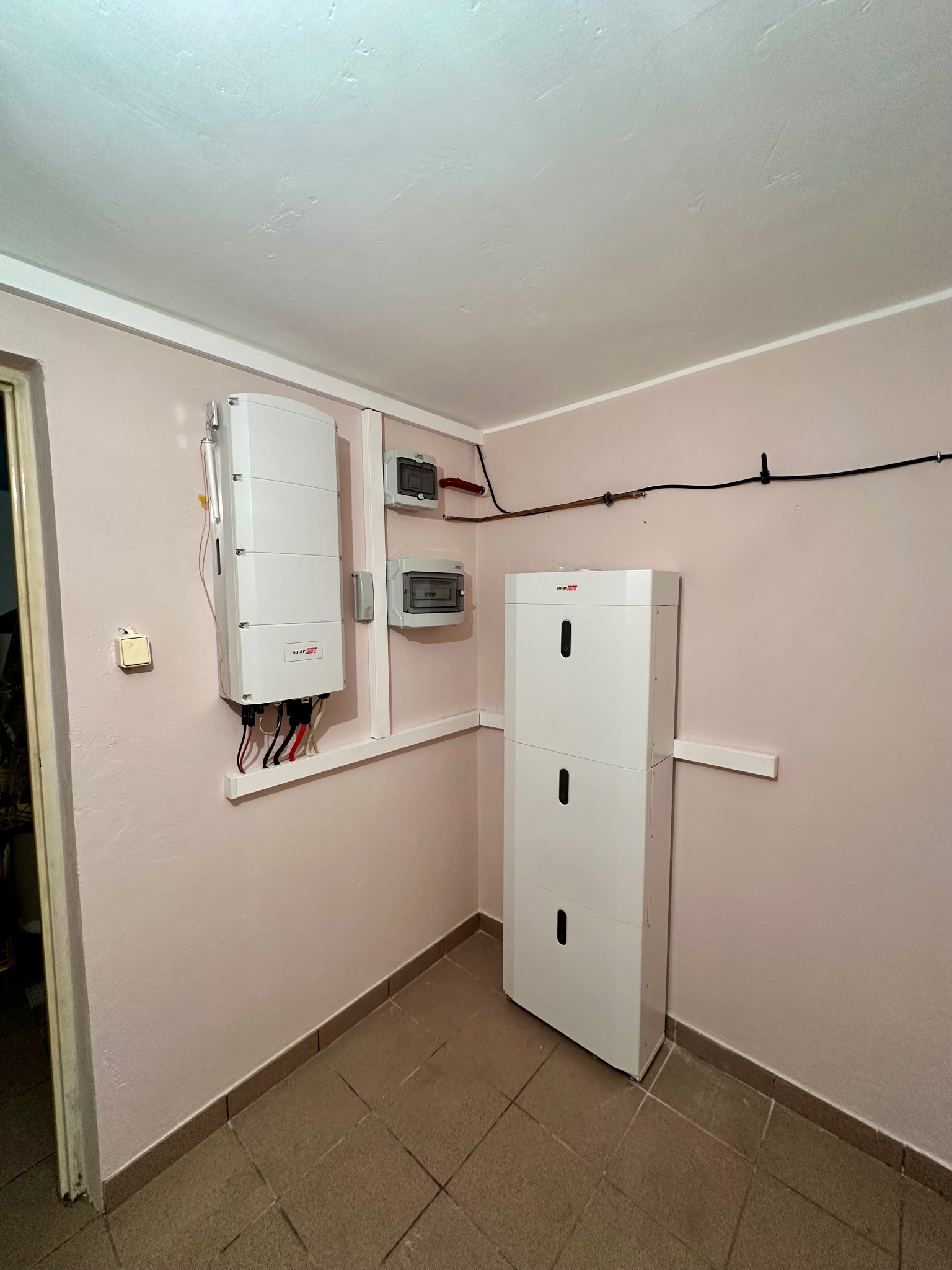 Wnętrze pomieszczenia z zamontowanym magazynem energii SolarEdge, inwerterem i rozdzielnicą elektryczną na ścianie w kolorze pudrowego różu. Widoczne kable i przewody.