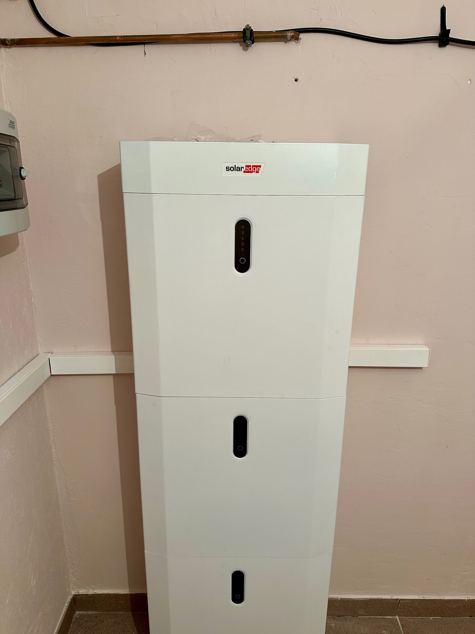Biały, trzypoziomowy magazyn energii SolarEdge na tle różowej ściany, widoczne przewody i instalacja elektryczna. Układ pionowy, oświetlenie naturalne.