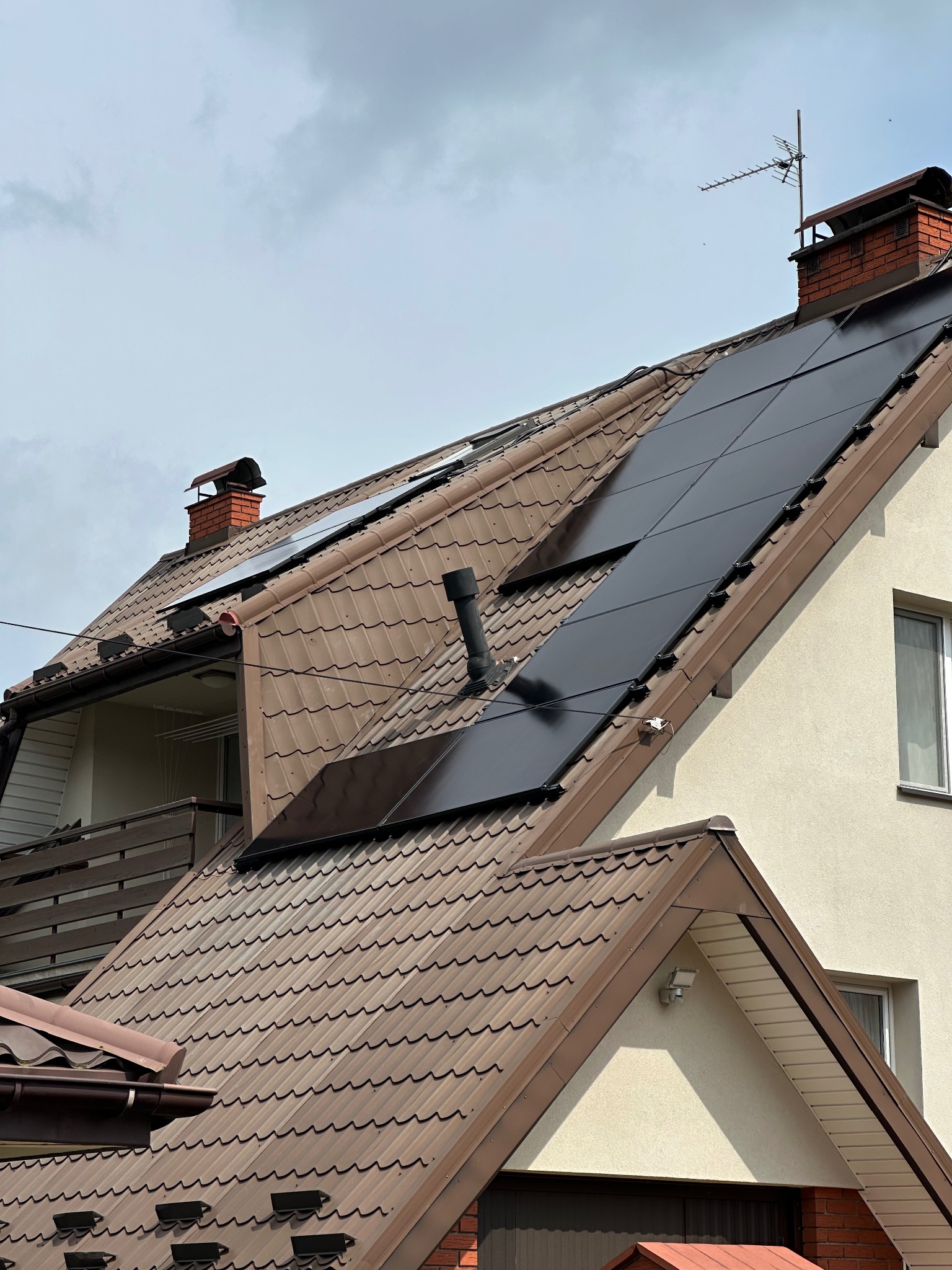 Brązowy dach z czarnymi panelami fotowoltaicznymi, komin z anteną. Ujęcie z dołu, częściowo widoczna elewacja domu w kolorze beżowym.