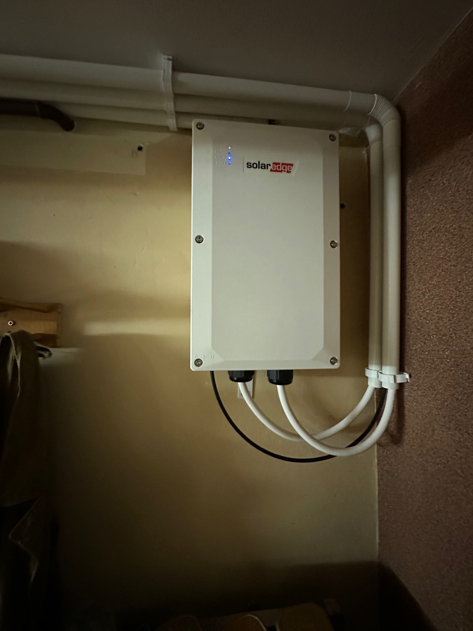 Biała skrzynka SolarEdge z podłączonymi kablami na ścianie. Widoczne rury instalacyjne przy suficie i w rogu pomieszczenia. Niebieskie diody sygnalizacyjne.