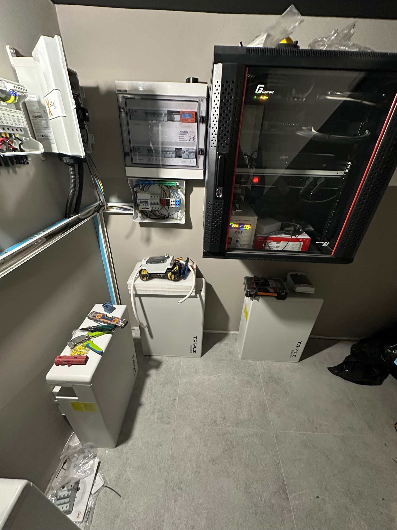 Instalacja magazynu energii Triple Power z rozdzielnicą i szafą rack. Widoczne narzędzia i elementy instalacji. Szara podłoga, neutralne oświetlenie.