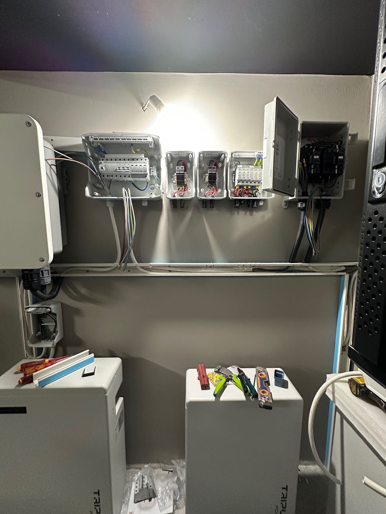 Rozdzielnia elektryczna z przewodami, skrzynkami i narzędziami na tle szarej ściany. Widoczne elementy instalacji i narzędzia pracy elektryka.