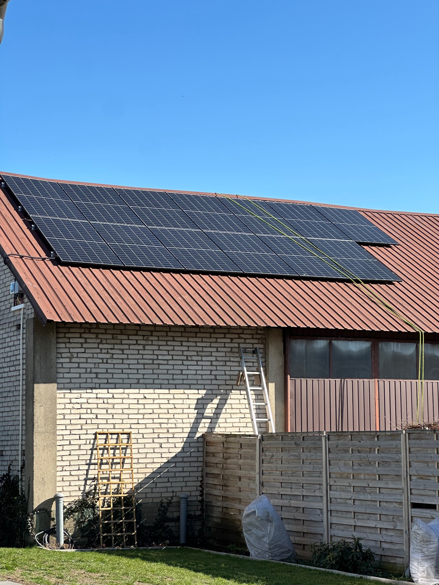 Panele słoneczne na dachu budynku z cegły, drabina oparta o ścianę. Widoczne przewody i elementy montażowe. Błękitne niebo w tle, słoneczny dzień.