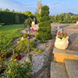 Urokliwy ogr&oacute;d z ścieżką z kostki, ozdobnymi roślinami, kamieniami, lampą i donicą w kształcie kwiatu na schodach. Zadbane trawniki i drzewa w tle.