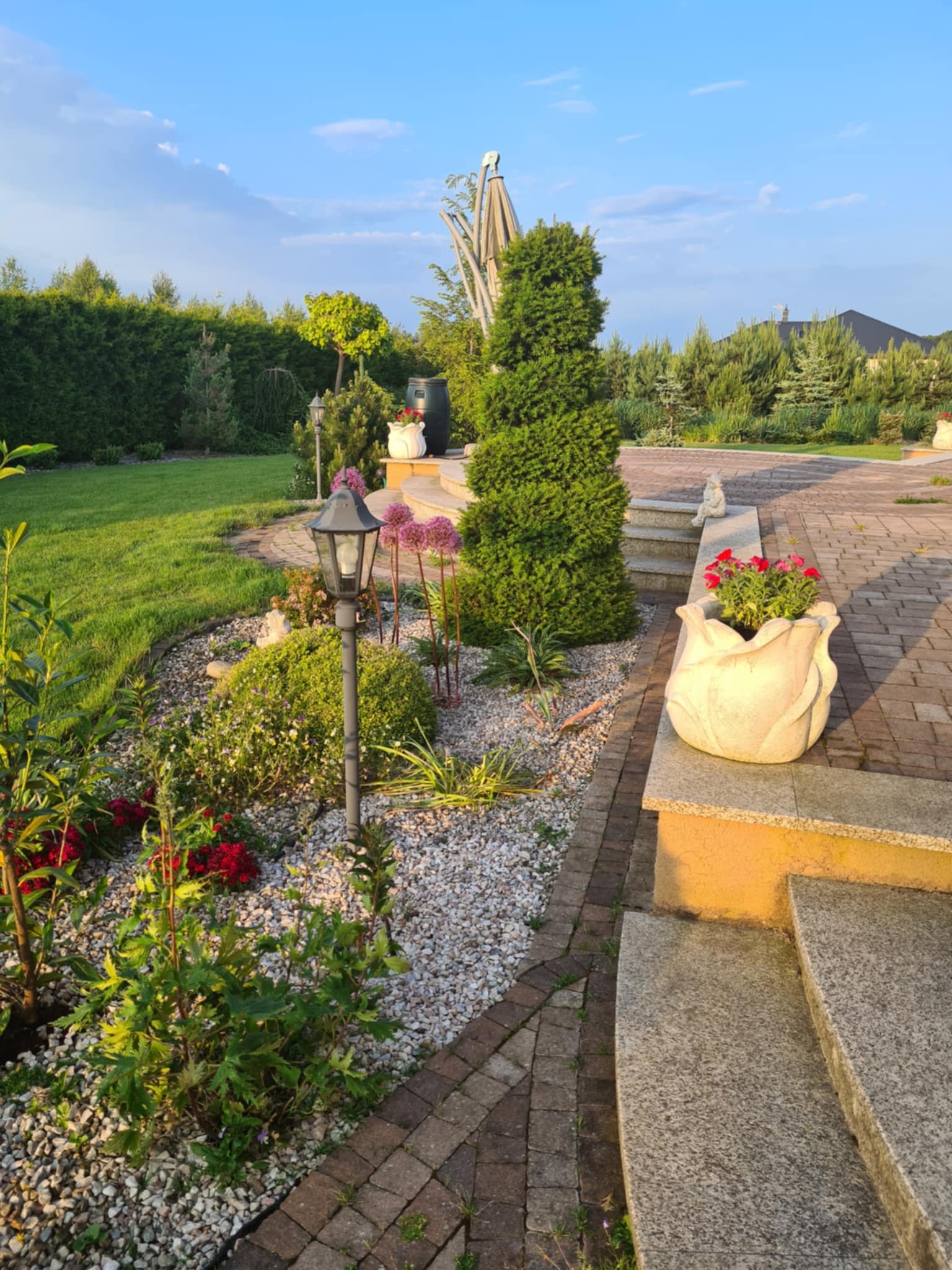 Urokliwy ogród z ścieżką z kostki, ozdobnymi roślinami, kamieniami, lampą i donicą w kształcie kwiatu na schodach. Zadbane trawniki i drzewa w tle.