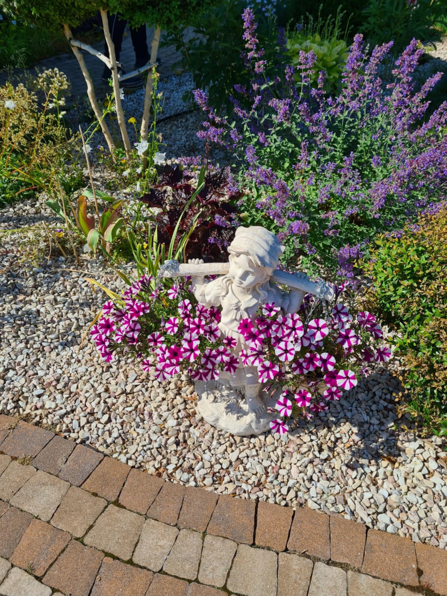 Ogród z rzeźbą chłopca otoczoną kwitnącymi petuniami w kolorze fioletowo-białym, na tle kamyczków i roślinności. Dekoracja ogrodu.