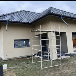 Dom jednorodzinny w trakcie prac wykończeniowych elewacji. Widoczne rusztowanie i zabezpieczone okna folią.
