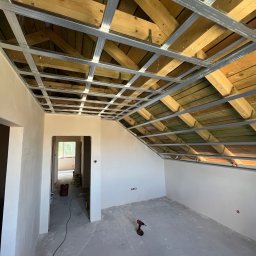 Jakub Owczarek Usługi Budowlane - Montaż stelaża sufitu podwieszanego z płyt karton-gips w pomieszczeniu ze świeżo wygładzonymi ścianami. Widoczna wkrętarka na podłodze.