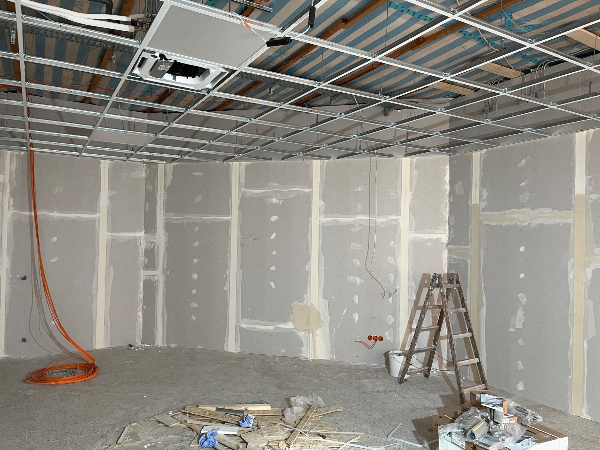 Montaż płyt gipsowo-kartonowych na ścianach i suficie. Widoczna konstrukcja metalowa sufitu podwieszanego i instalacja elektryczna.  Na podłodze leżą narzędzia i materiały budowlane.