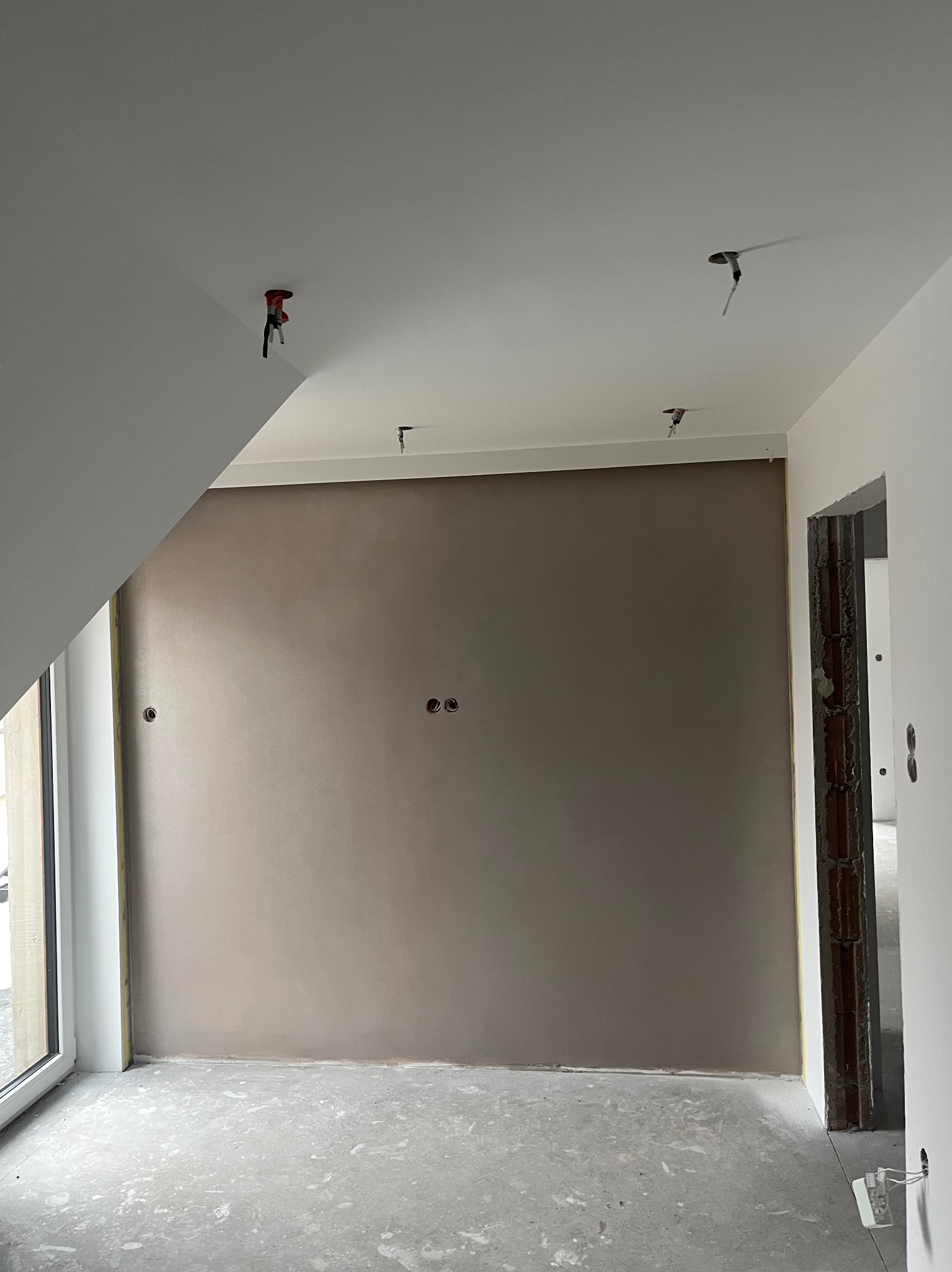 Wnętrze w trakcie wykańczania: gładka, beżowa ściana z widocznymi puszkami elektrycznymi, surowa podłoga, instalacja oświetleniowa w suficie. Skos dachu.