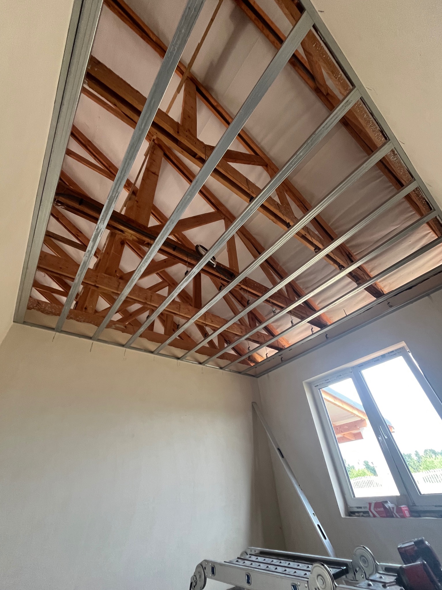 Montaż metalowej konstrukcji sufitu podwieszanego na drewnianej więźbie dachowej. Widoczne profile i elementy mocujące. W tle okno i drabina.