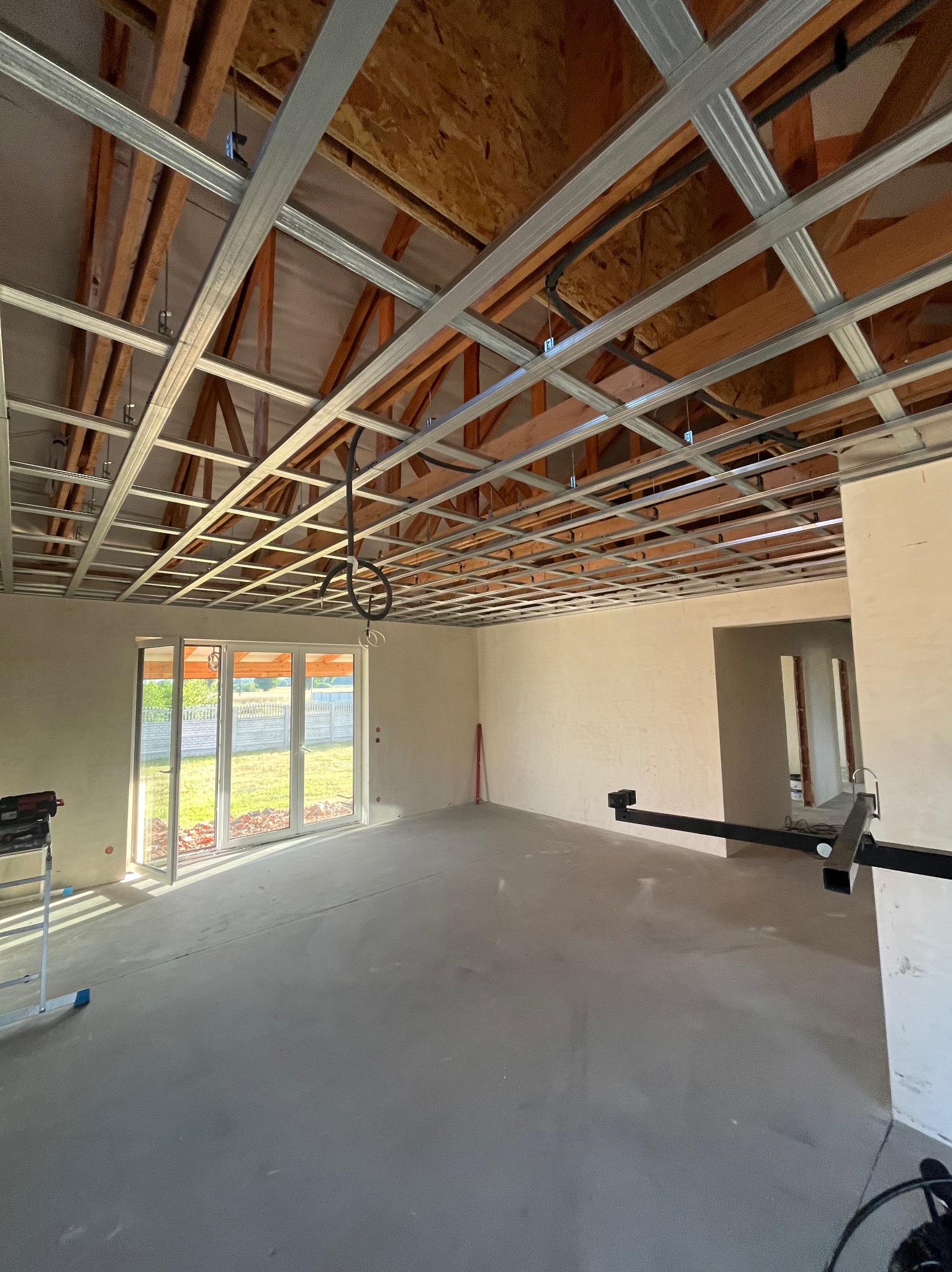 Montaż metalowej konstrukcji sufitu podwieszanego w jasnym wnętrzu z dużym oknem. Widoczne drewniane elementy więźby dachowej i narzędzia pracy.