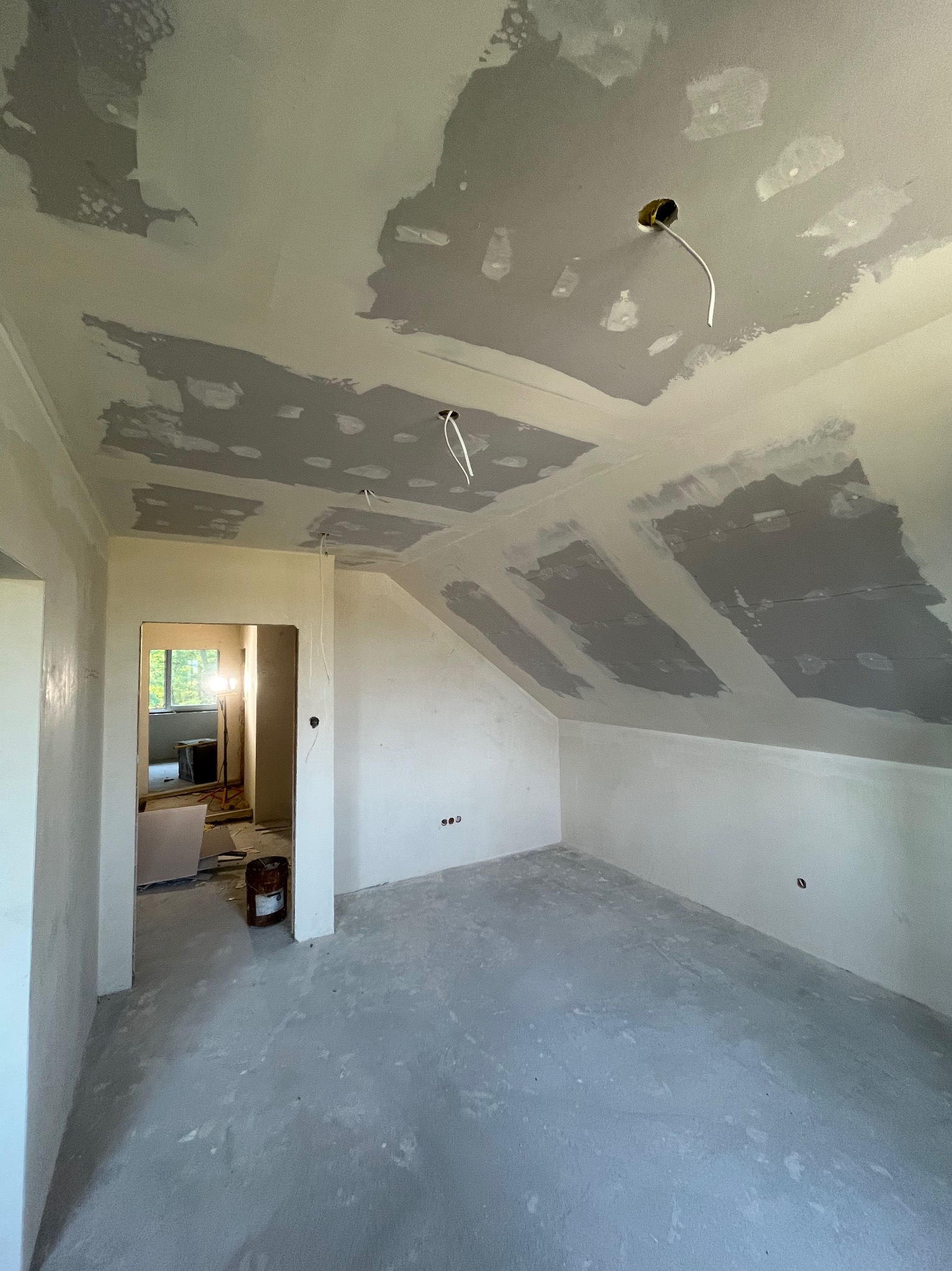 Pokój na poddaszu w trakcie prac wykończeniowych: ściany i sufit częściowo pokryte gładzią, widoczne kable elektryczne. W tle otwarte przejście do innego pomieszczenia.