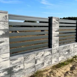 Nowoczesne ogrodzenie z szarych, betonowych bloczków o wzorze marmuru i poziomych, metalowych przęseł w odcieniu grafitu na tle błękitnego nieba.