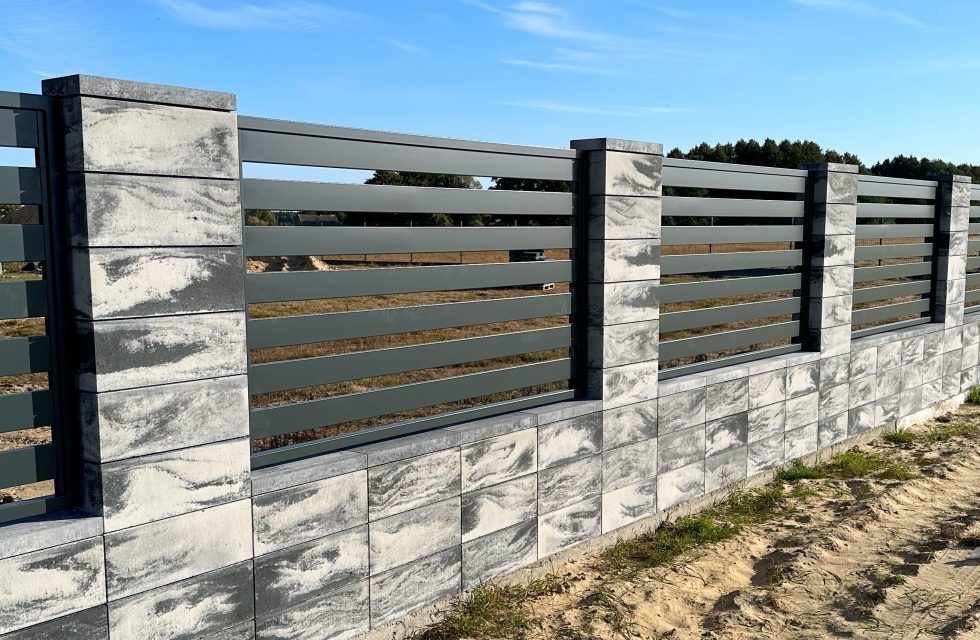 Nowoczesne ogrodzenie z szarych, betonowych bloczków o wzorze marmuru i poziomych, metalowych przęseł w odcieniu grafitu na tle błękitnego nieba.