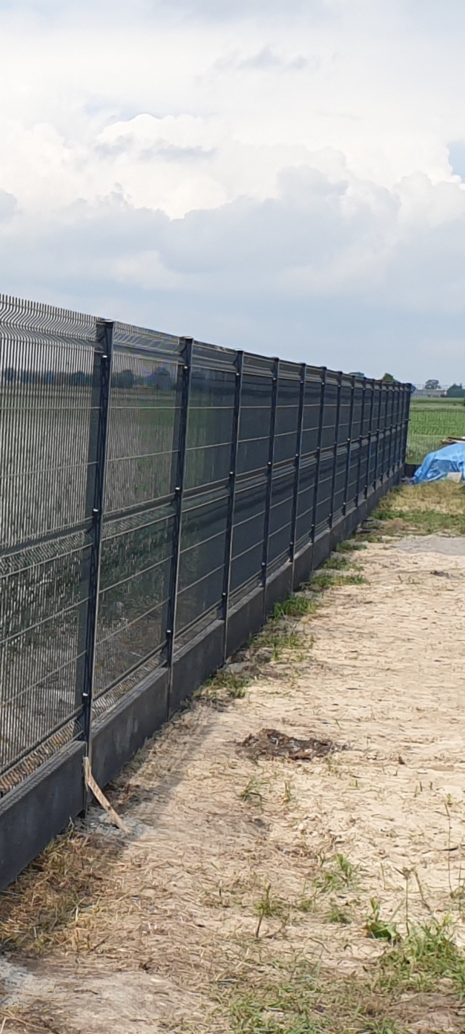 Długi, panelowy, grafitowy płot z podmurówką na piaszczystym terenie, w tle pola i błękitne niebo z chmurami. Widok z boku, prezentacja montażu ogrodzenia panelowego.