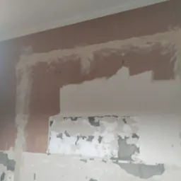 Ściana w trakcie remontu: częściowo otynkowana, z widocznymi śladami po usuniętych płytkach i starych warstwach farby. Fragmenty brązowej farby widoczne w górnej części.