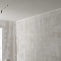 Ściana po nałożeniu gładzi, przygotowana do malowania. Widoczne przewody elektryczne wystające z sufitu. Ściany w jasnych, neutralnych odcieniach szarości i bieli.