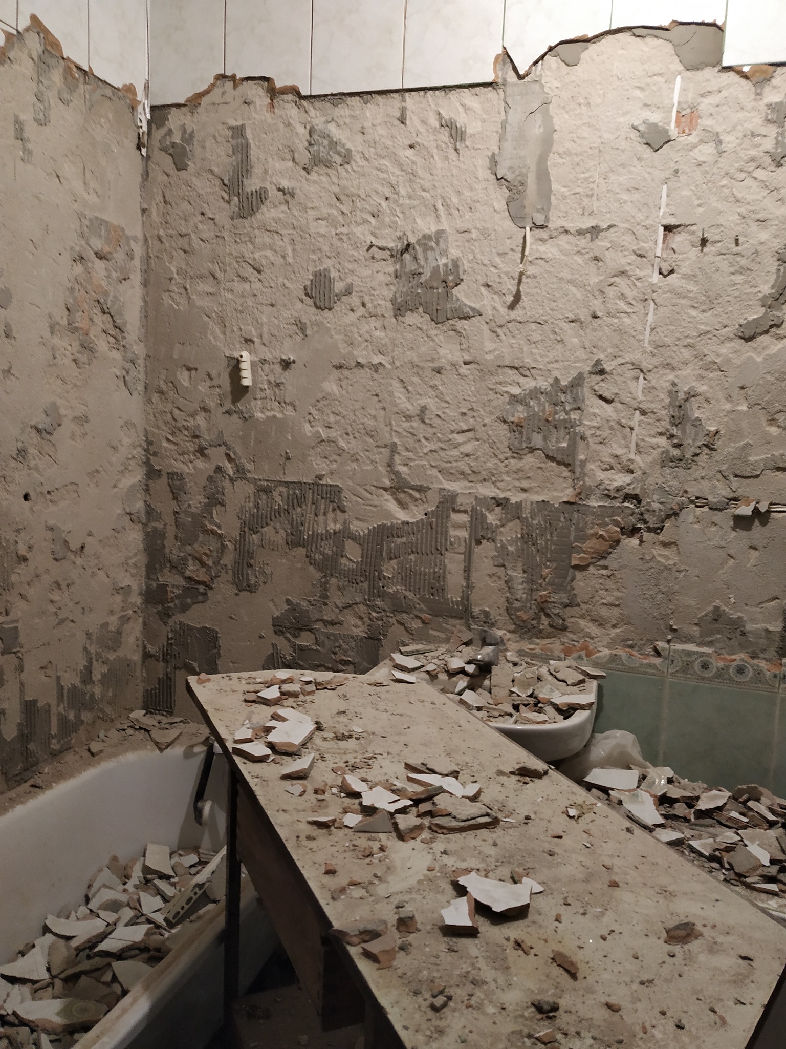 Zdemontowane płytki w łazience, gruz na prowizorycznym stole i w wannie. Ściany z pozostałościami kleju i fragmentami płytek. Widoczne stare instalacje.